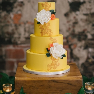 Caribbean Inspired, 4- Tiered Round Wedding Cake on Wooden Table with Yellow Fondant and Gold Design Detailing Accented with White Flowers