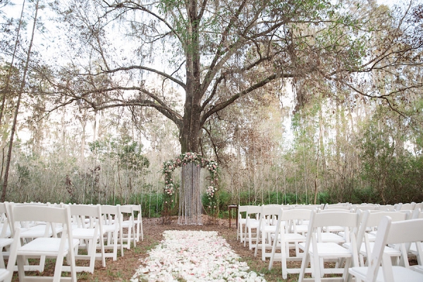 Rustic Outdoor Wedding Ceremony with Light Pink and White Floral Archway under Tall Pines with White Resin Folding Chairs and Rose Petal Aisle 
